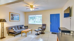 A living room with a sofa, coffee table, and armchair, a ceiling fan, a window with blinds, a blue door, and a flat-screen TV mounted on the wall. - Pet-friendly Hallandale Beach Condo Near Boardwalk (Hallandale Beach)