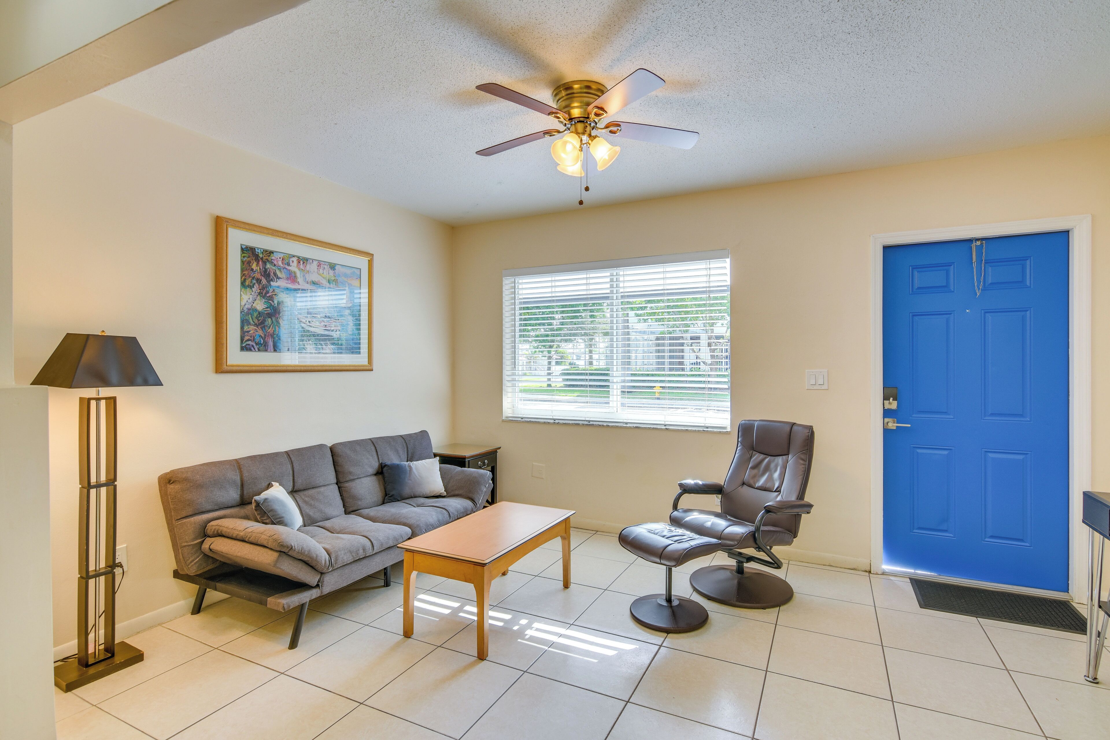 A living room with a sofa, coffee table, and armchair, a ceiling fan, a blue door, and a window with blinds.