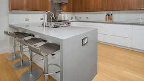 Cozinha moderna com armários de madeira, ilha central com banquetas, eletrodomésticos de aço inoxidável e painel de azulejos.