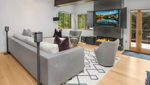 Uma sala de estar moderna com um sofá cinzento, lareira, televisão de tela plana e janelas amplas.