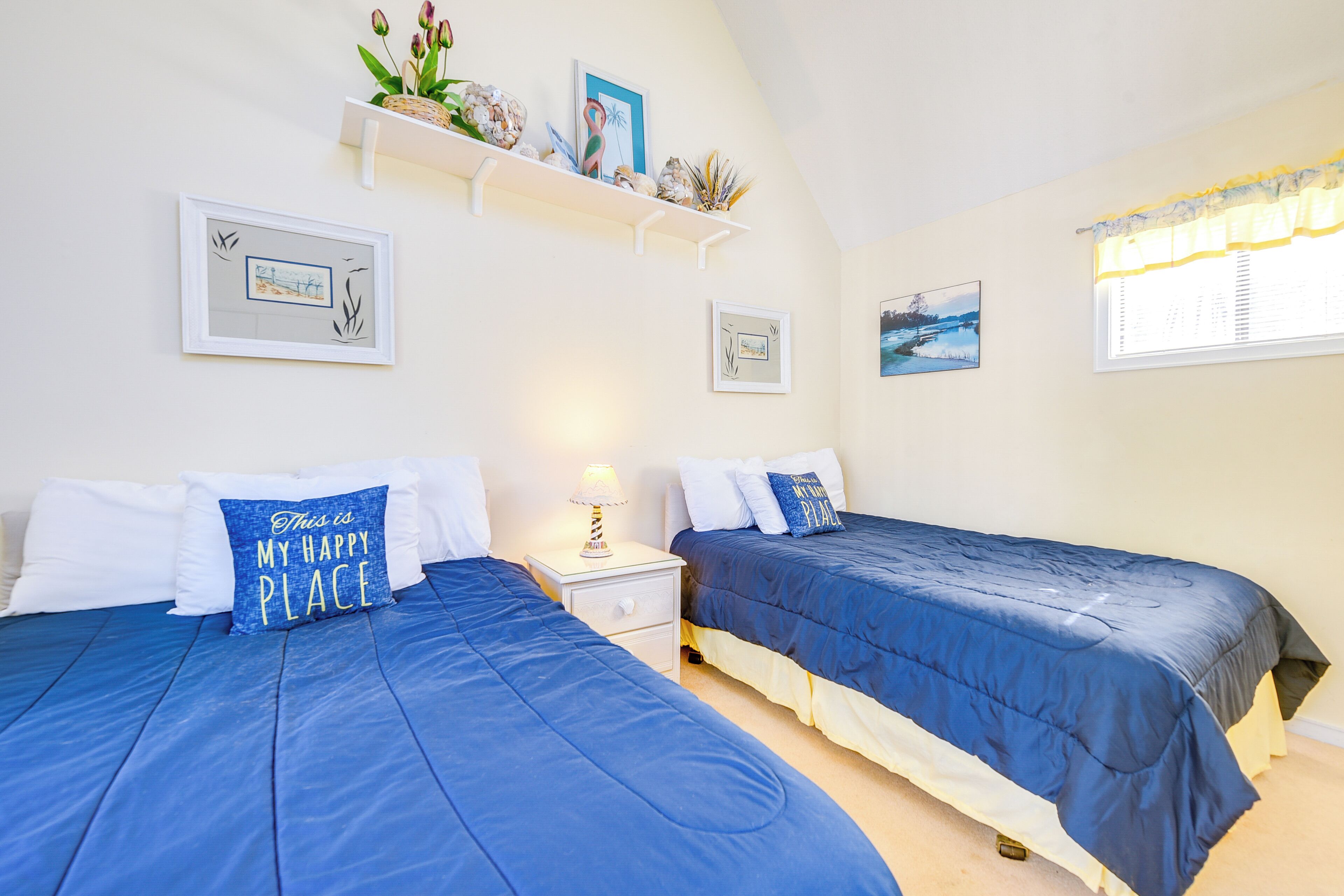 A bedroom with two beds, each with blue bedding and white pillows. There are wall-mounted shelves with decorative items and framed pictures.