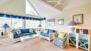 A bright living room with a blue sofa, white armchairs, and a patterned armchair. There are two lamps, a bookshelf, and a ceiling fan.