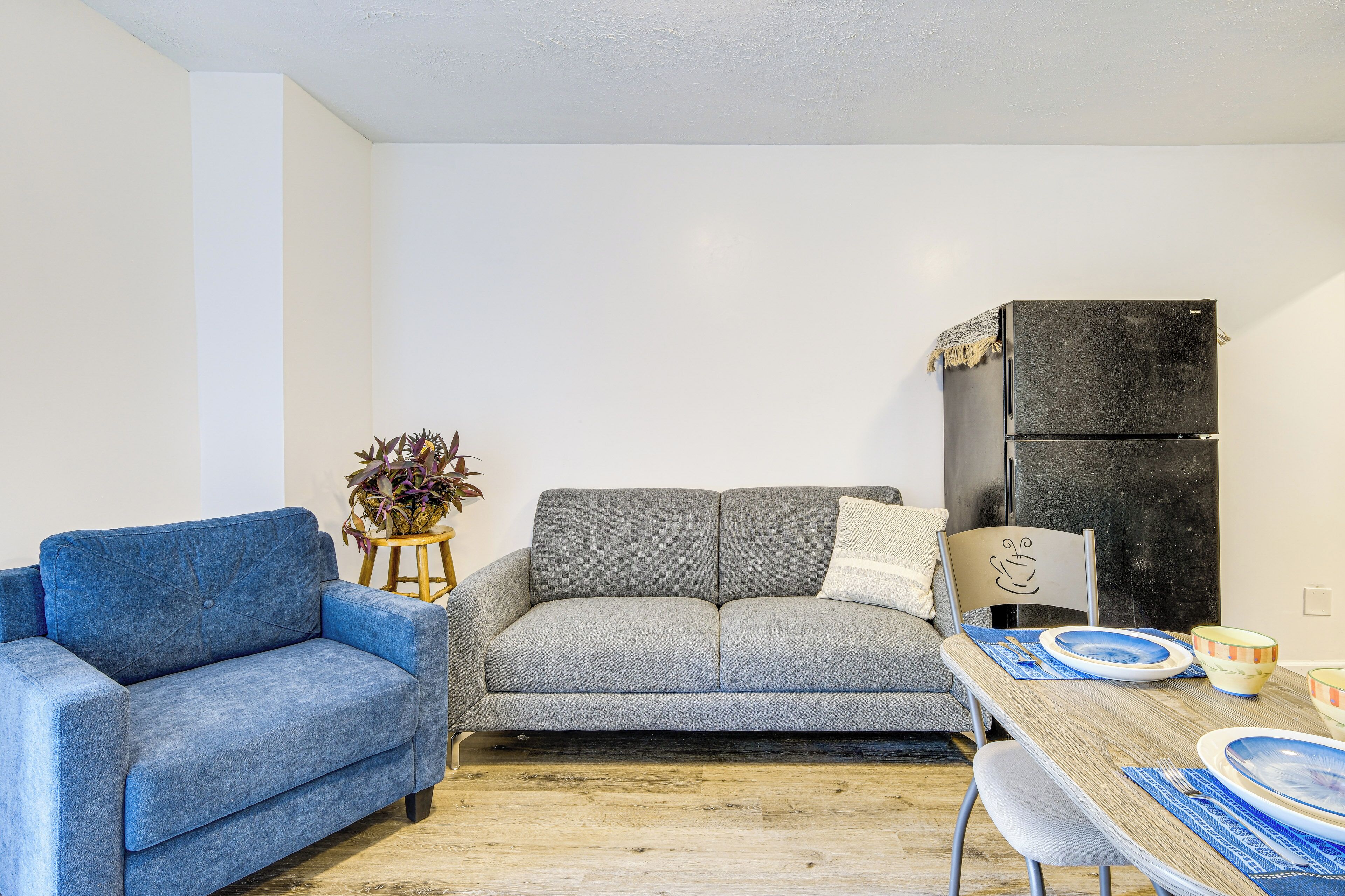 A living room with a blue sofa, a grey sofa, a wooden dining table with blue and white plates, and a black refrigerator.