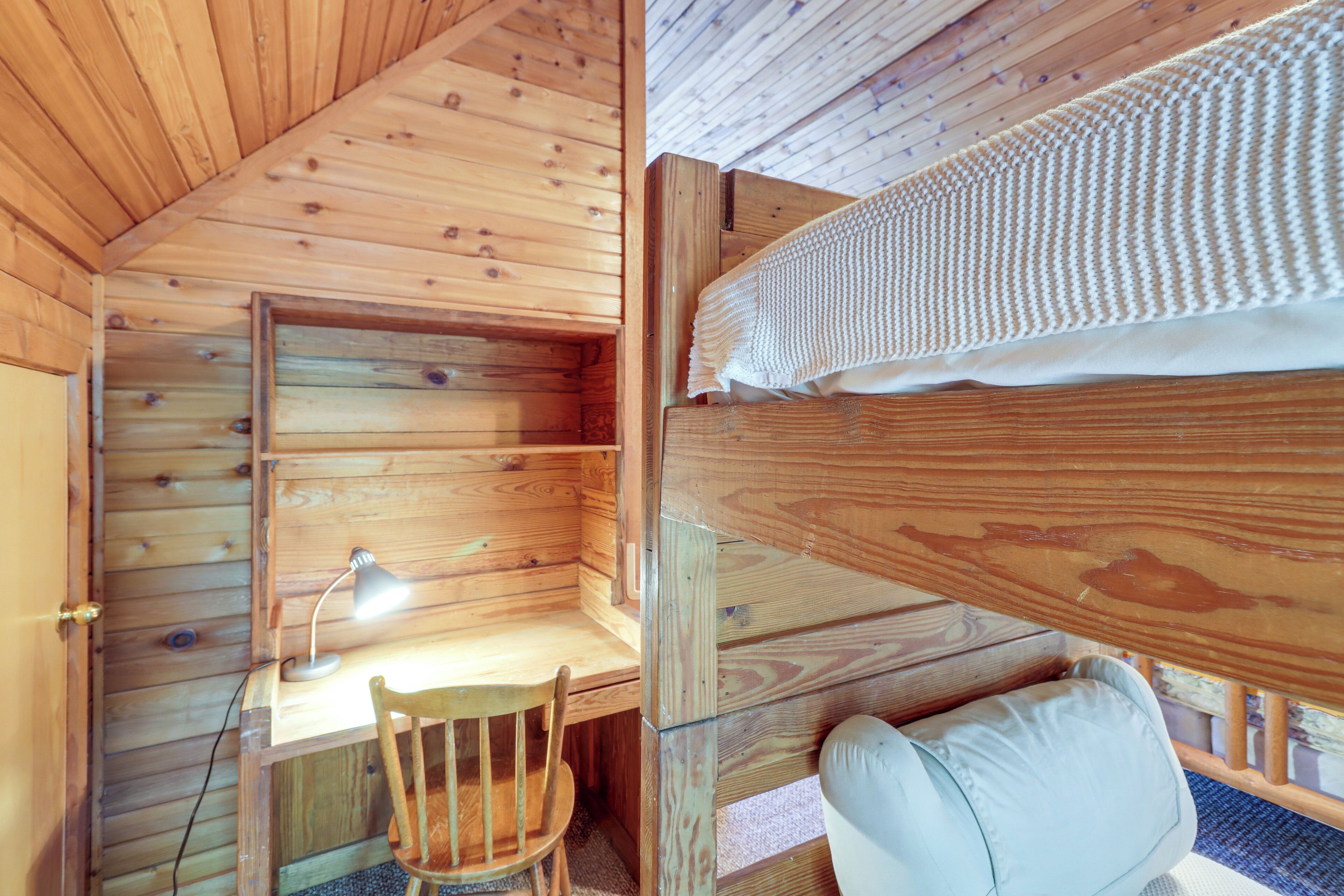 A wooden cabin interior with a bunk bed, a desk with a lamp, and a chair.