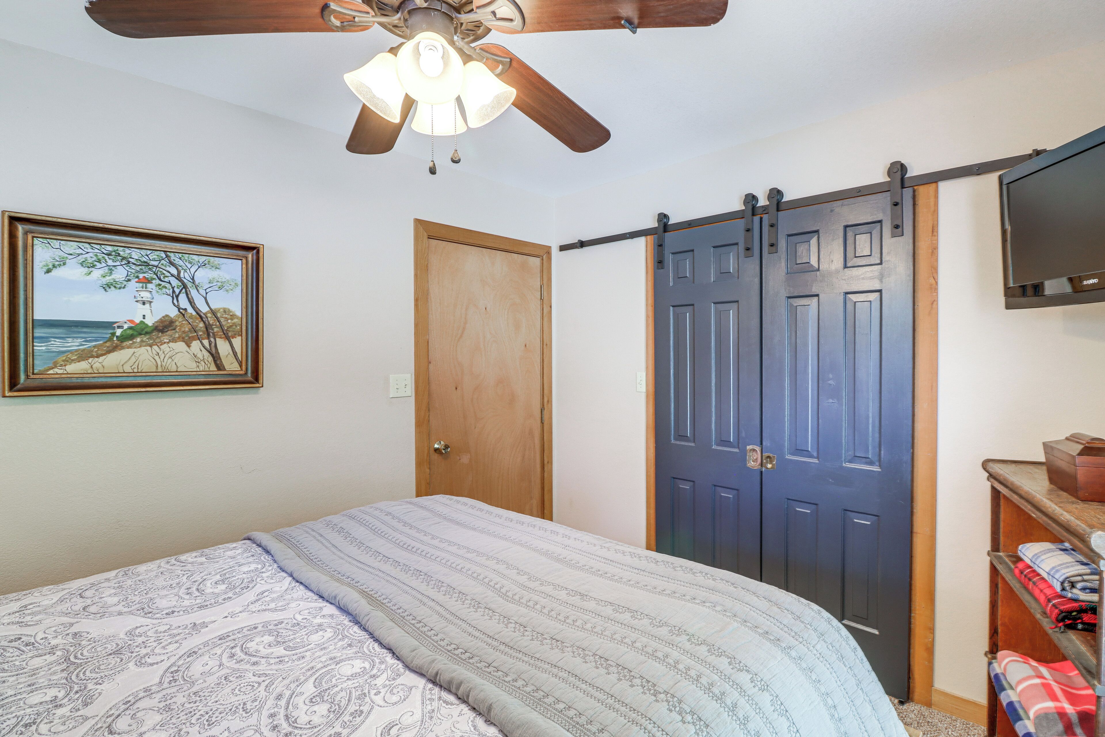 A bedroom with a bed, a ceiling fan, a wooden door, a sliding barn door, a TV mounted on the wall, and a framed picture on the wall.