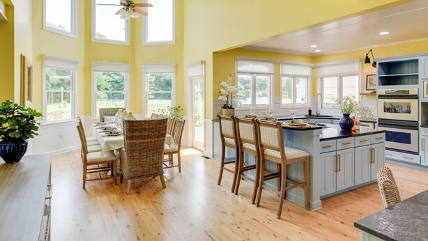 A bright kitchen with large windows, a dining area with wicker chairs, and a central island with bar stools.