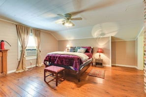 A bedroom with a wooden floor, a bed with a purple bedspread, a ceiling fan, a nightstand with a lamp, and a window with curtains.