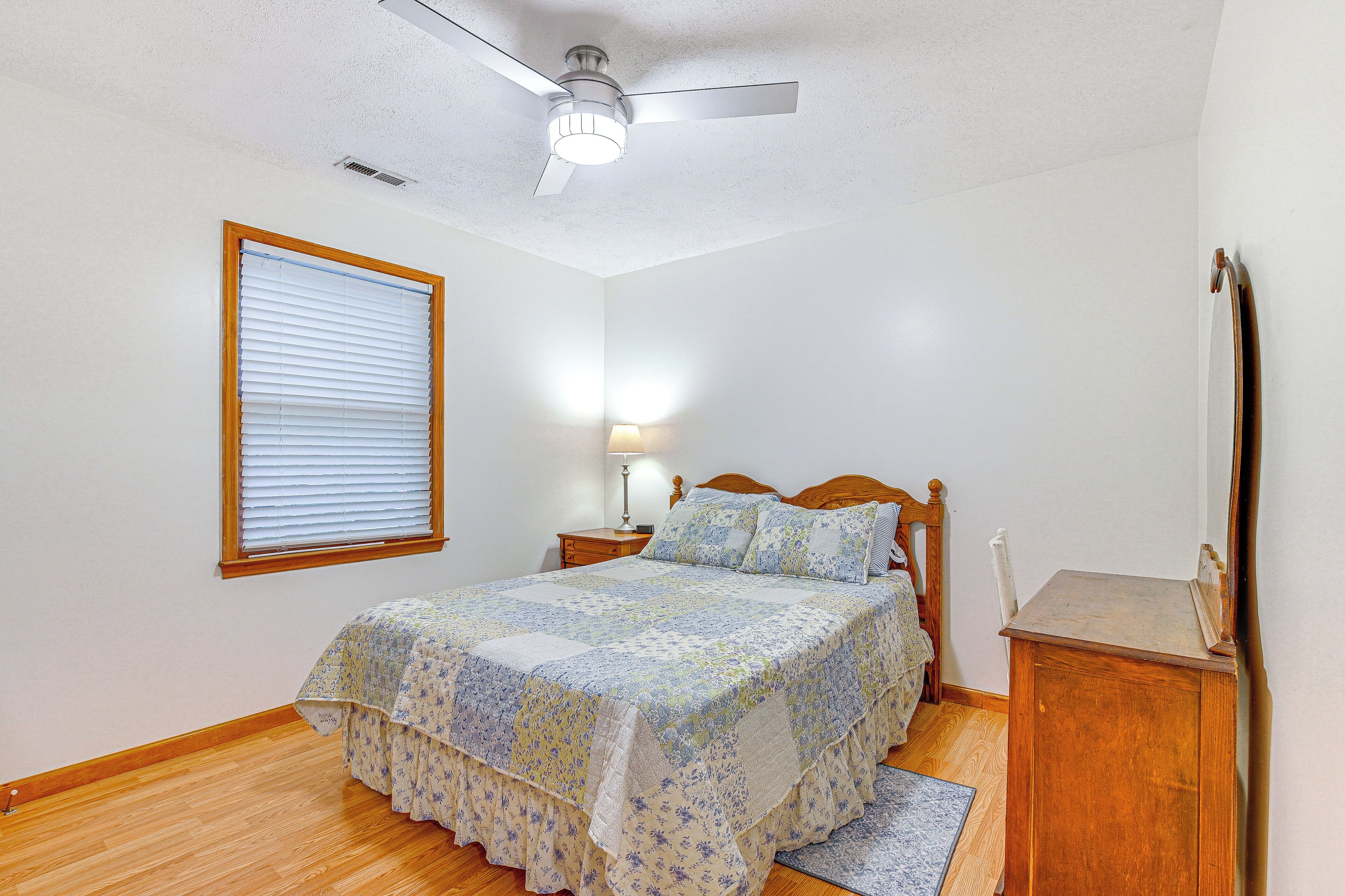 A bedroom with a wooden floor, a bed with a floral bedspread, a nightstand with a lamp, a wooden dresser, and a ceiling fan.