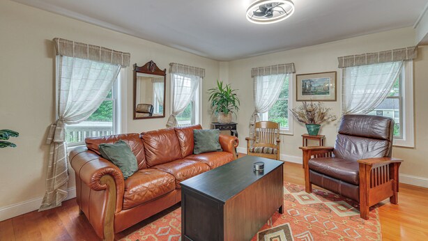 A living room with a leather sofa, a wooden coffee table, and a leather armchair. There are windows with sheer curtains, a ceiling fan, and a framed picture on the wall. - Quintessential New England Home w/ Patio & Grill! (Putnam)