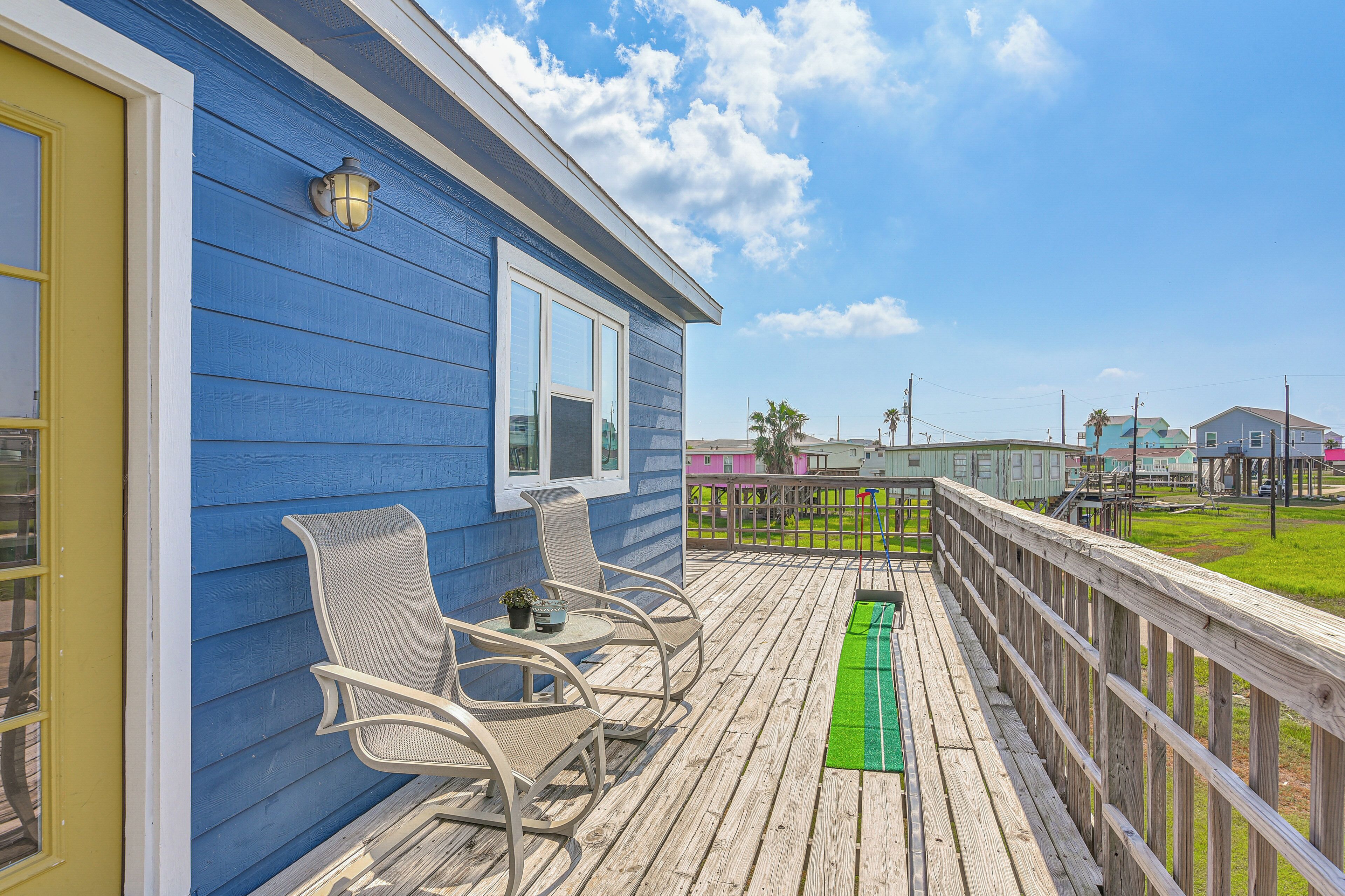 Een houten terras met twee stoelen en een tafel, een blauw huis met een gele deur, en een uitzicht op andere huizen op de achtergrond.