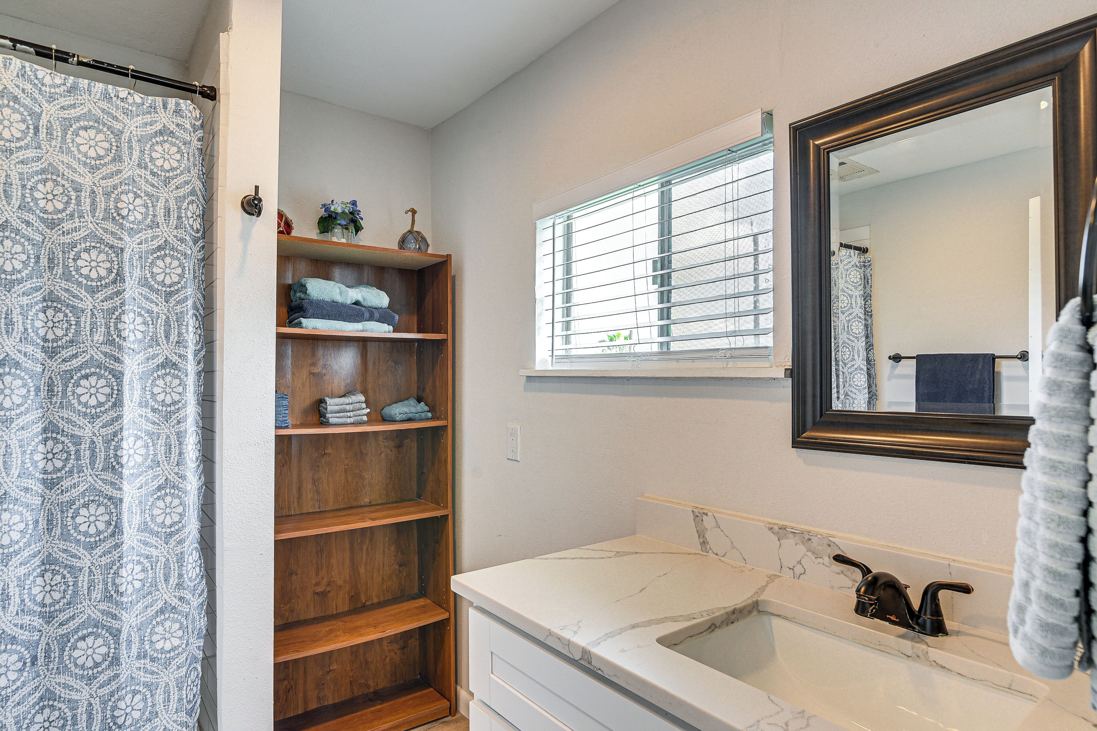 A bathroom with a shower curtain, a wooden shelf with towels, a window with blinds, a sink with a mirror, and a towel rack.