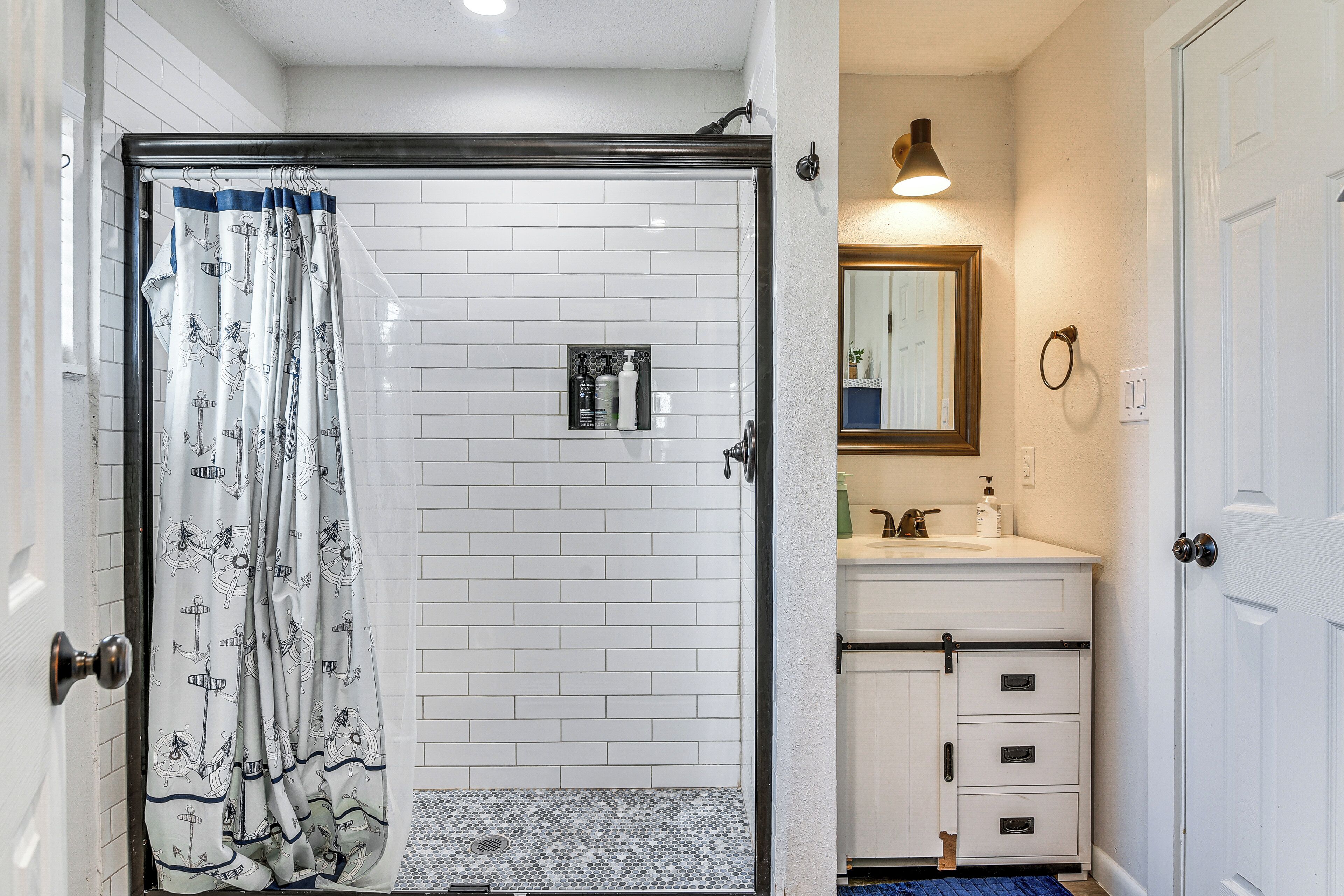 A bathroom with a shower, white tiled walls, a sink with a mirror, and a door.