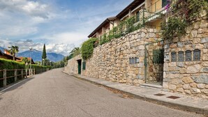 A stone building with balconies, a paved street, and mountains in the background. - Holiday Home 'Casa Di Tino' with Lake View, Wi-Fi and Air Conditioning (Brenzone sul Garda)
