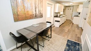 A modern kitchen with white cabinets, a dining area with a table and chairs, and a large painting on the wall. - 3 blocks to Notre Dame, EV Charger, BBQ & Fire Pit! (1707) (South Bend)