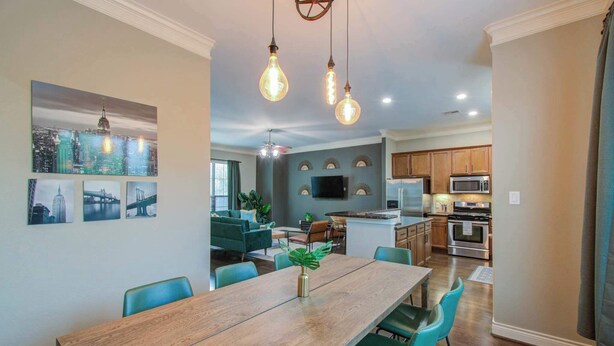 Un vaste espace de salle à manger avec une table en bois et des chaises de couleur turquoise, une cuisine équipée d’appareils électroménagers modernes et un tableau représentant un paysage urbain accroché au mur.