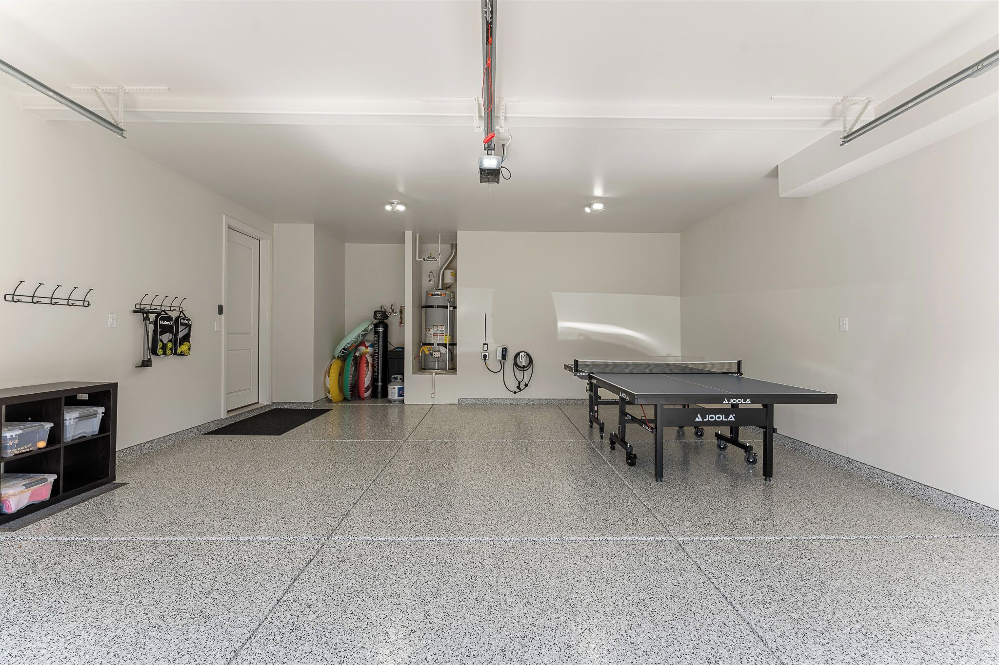 Ein sauberes, modernes Zimmer mit einem Tischtennistisch, einem schwarzen Regalelement mit Aufbewahrungsboxen und einer Tür zu einem weiteren Bereich.