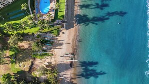 Vue aérienne d’un complexe hôtelier en bord de mer, avec une piscine, des palmiers et des parasols de plage.
