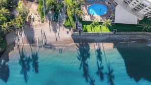 Vue aérienne d’un complexe hôtelier de bord de mer, avec une piscine, des palmiers et une plage de sable.