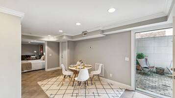 A dining area with a round table and white chairs, a kitchen in the background, and a view of the outside through a glass door.