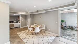 Un coin repas avec une table ronde et des chaises blanches, une cuisine en arrière-plan et une vue sur l’extérieur par une porte vitrée.