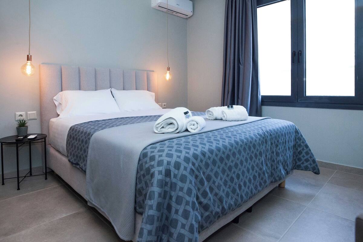 A neatly made bed with a patterned bedspread, a bedside table with a lamp, and a window with curtains.