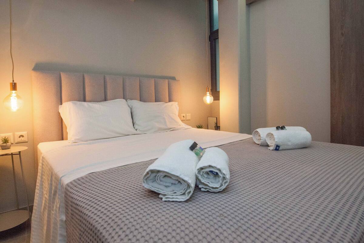 A neatly made bed with white linens and rolled towels, a bedside table with a lamp, and a small potted plant.