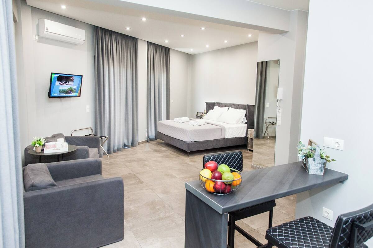 A modern hotel room with a bed, a desk with a fruit bowl, a sofa, and a TV on the wall.