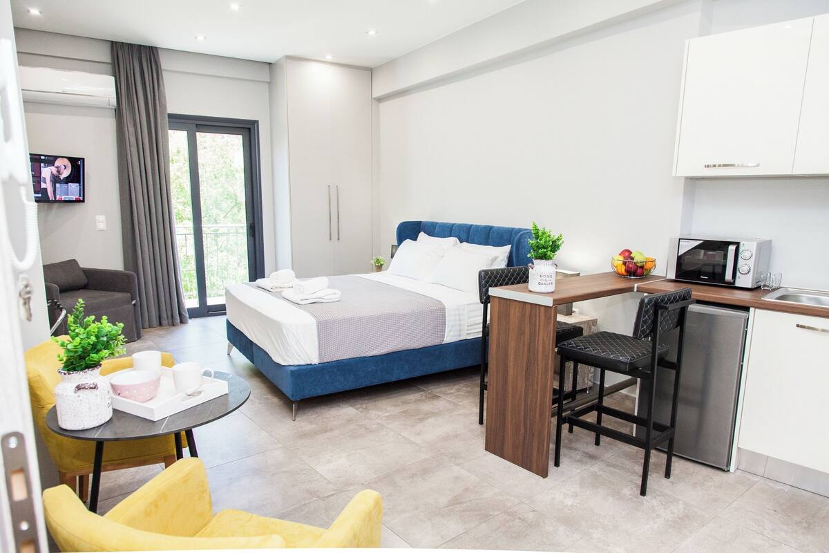 A modern hotel room with a bed, a desk, a kitchenette, and a seating area.