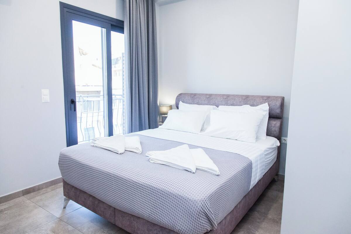 A neatly made bed with white linens and a grey patterned bedspread, a window with blue curtains, and a door leading outside.