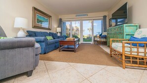 A living room with a grey sofa, a blue sofa, a wooden coffee table, and a wicker chair. There is a television mounted on the wall and a window with a view of palm trees.