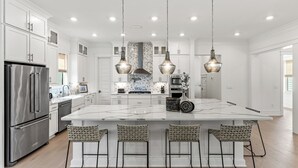 A modern kitchen with a large island, stainless steel appliances, hanging pendant lights, and bar stools. - Spacious New Home in Blue Mountain Beach (Santa Rosa Beach)