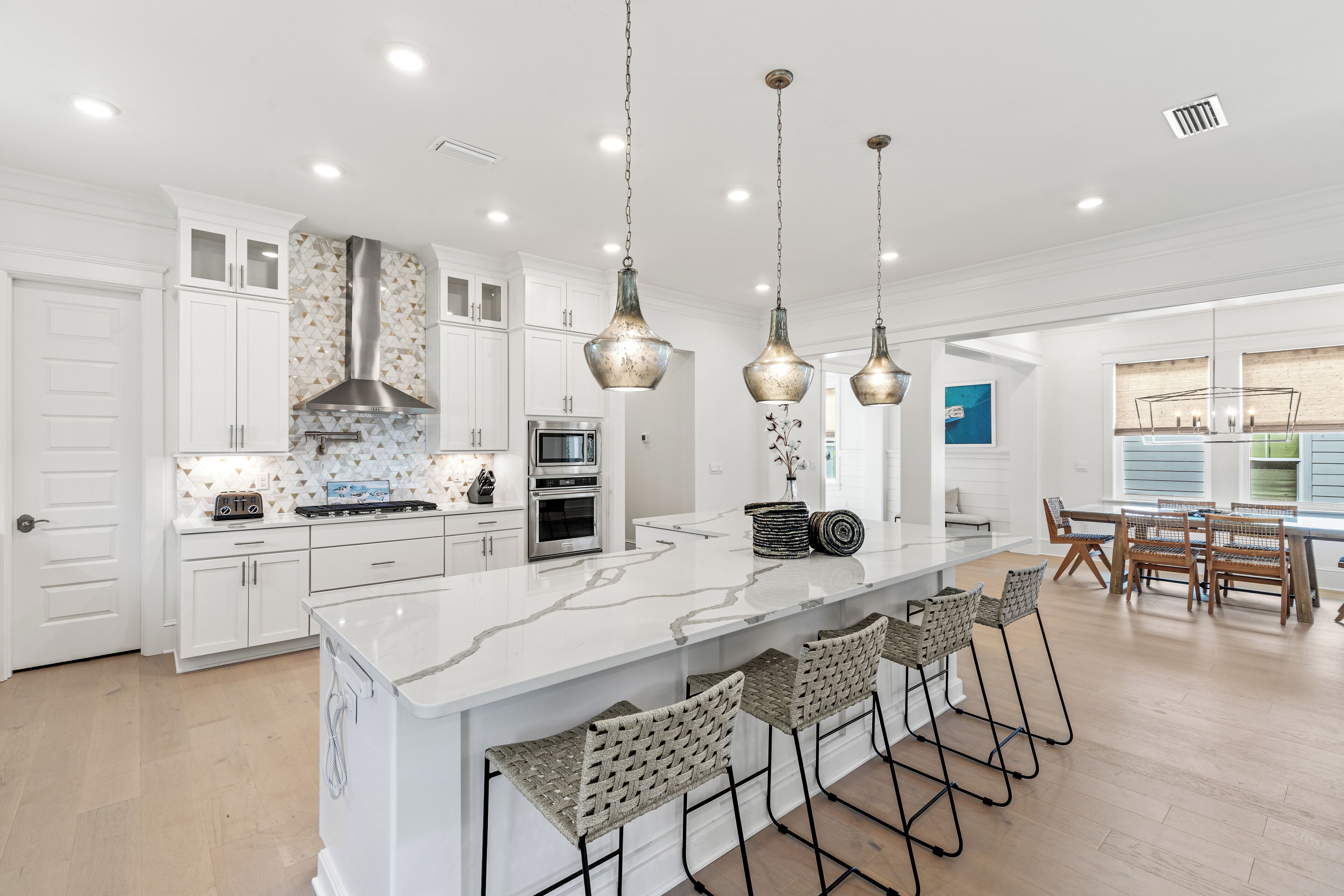 A modern kitchen with white cabinetry, a marble island, hanging pendant lights, and a dining area with wooden chairs and a table.