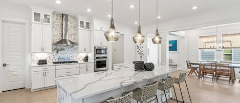 A modern kitchen with white cabinetry, a marble island, hanging pendant lights, and a dining area with wooden chairs and a table.