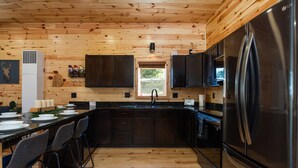 A modern kitchen with wooden walls, a stainless steel refrigerator, and a long countertop with bar stools. - Otsego Lake Access Cabin, Fire Pit, Backyard & BBQ (Gaylord)