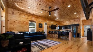 A spacious living area with a wooden ceiling, a blue sofa, a kitchen with modern appliances, and a dining area with bar stools. - Otsego Lake Access Cabin, Fire Pit, Backyard & BBQ (Gaylord)