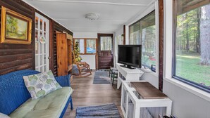 A cozy living room with a blue sofa, a television, and large windows offering a view of the outdoors. - Rustic Willow Retreat - Outdoor Adventures 2 Bedroom Cabin by RedAwning (Hazelhurst)