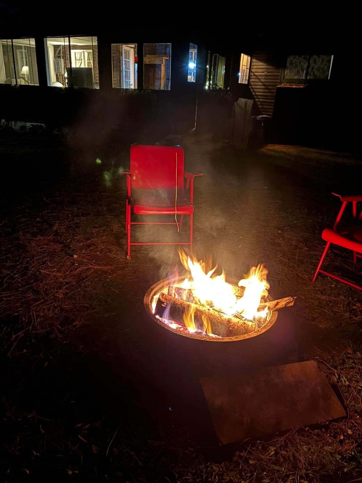 Escena nocturna al aire libre con un brasero, dos sillas rojas y un edificio con ventanas iluminadas al fondo.
