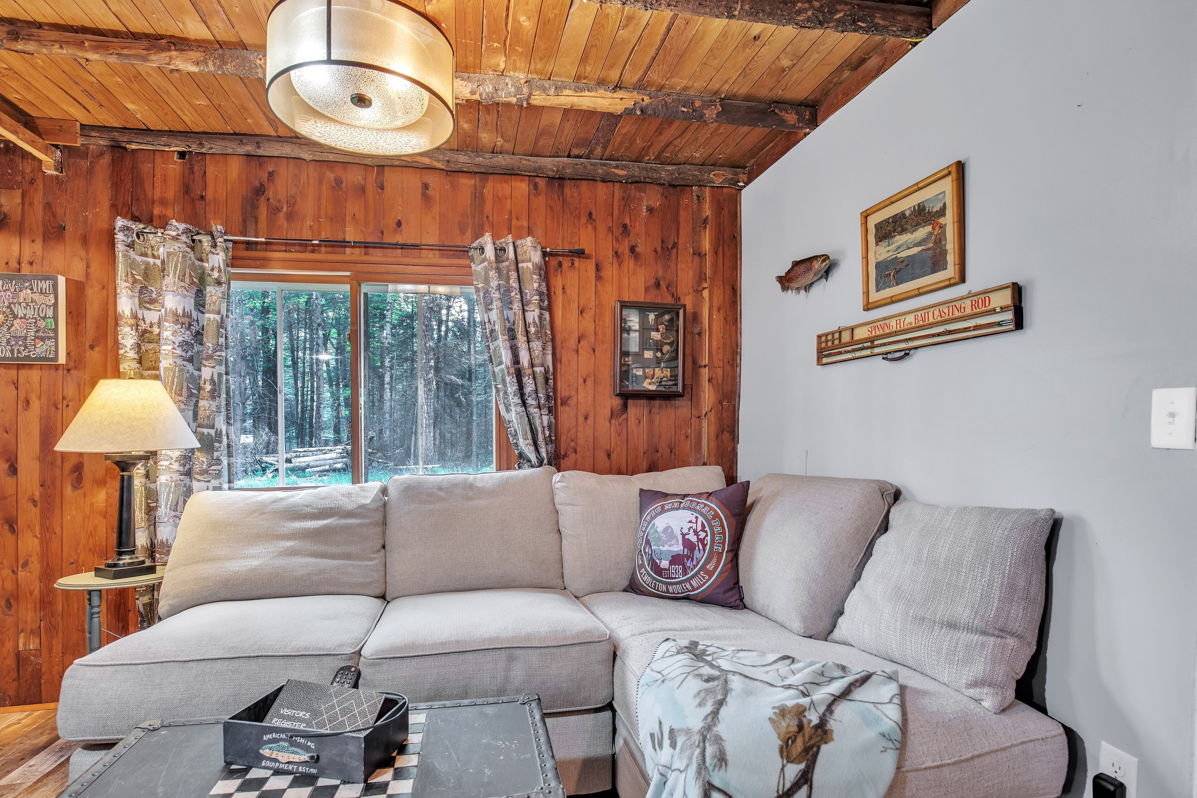 A cozy cabin interior with a sectional sofa, a lamp, a framed picture, and a window with curtains.