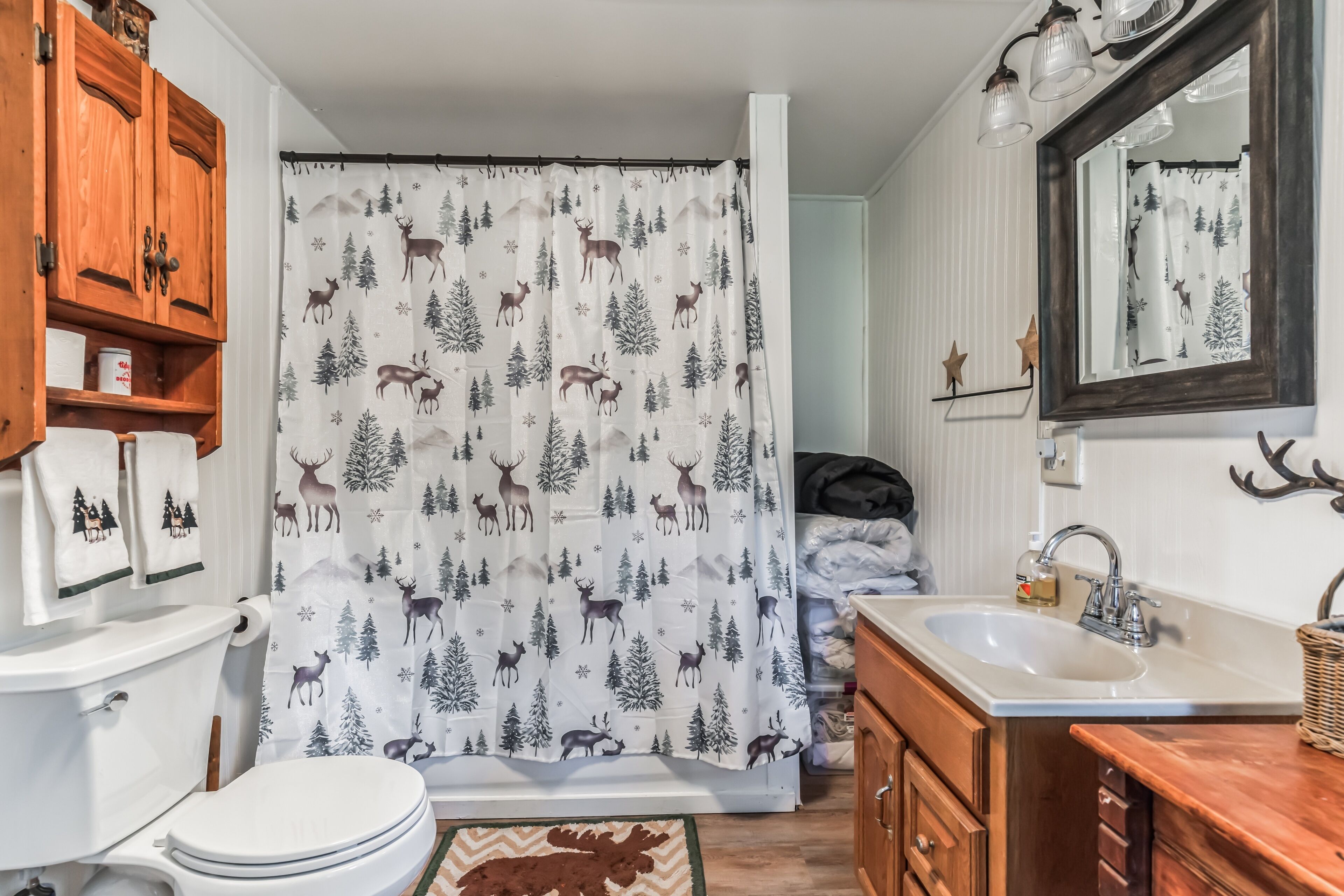 Un baño con inodoro, lavamanos, gabinete de madera y cortina de ducha con estampado de ciervo y árbol.