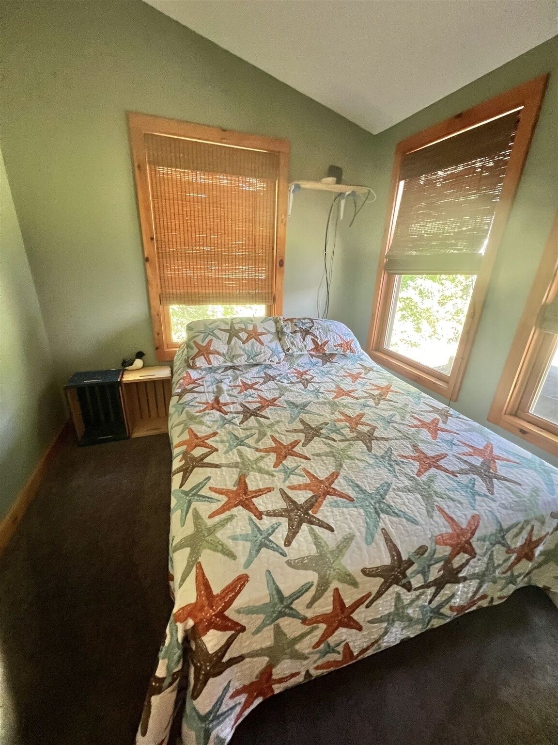 A bedroom with a bed, two windows with blinds, a small bedside table, and a wall-mounted heater.