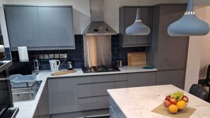 A modern kitchen with grey cabinets, a central island, a gas stove, and pendant lighting. - 4 Bedroom Luxury Home with Garden in East London (Ilford)