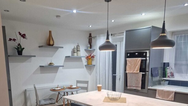 A modern kitchen with a central island, black bar stools, pendant lighting, and a variety of decorative items on shelves. - 4 Bedroom Luxury Home with Garden in East London (Ilford)