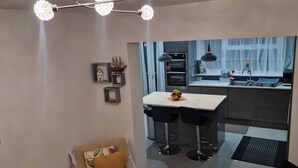 A modern kitchen with a central island, a dining area with bar stools, and a seating area with a cushioned chair. - 4 Bedroom Luxury Home with Garden in East London (Ilford)