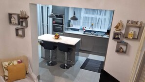A modern living room with a sofa and a coffee table, a dining area with a table and chairs, and a kitchen with a sink and oven. - 4 Bedroom Luxury Home with Garden in East London (Ilford)