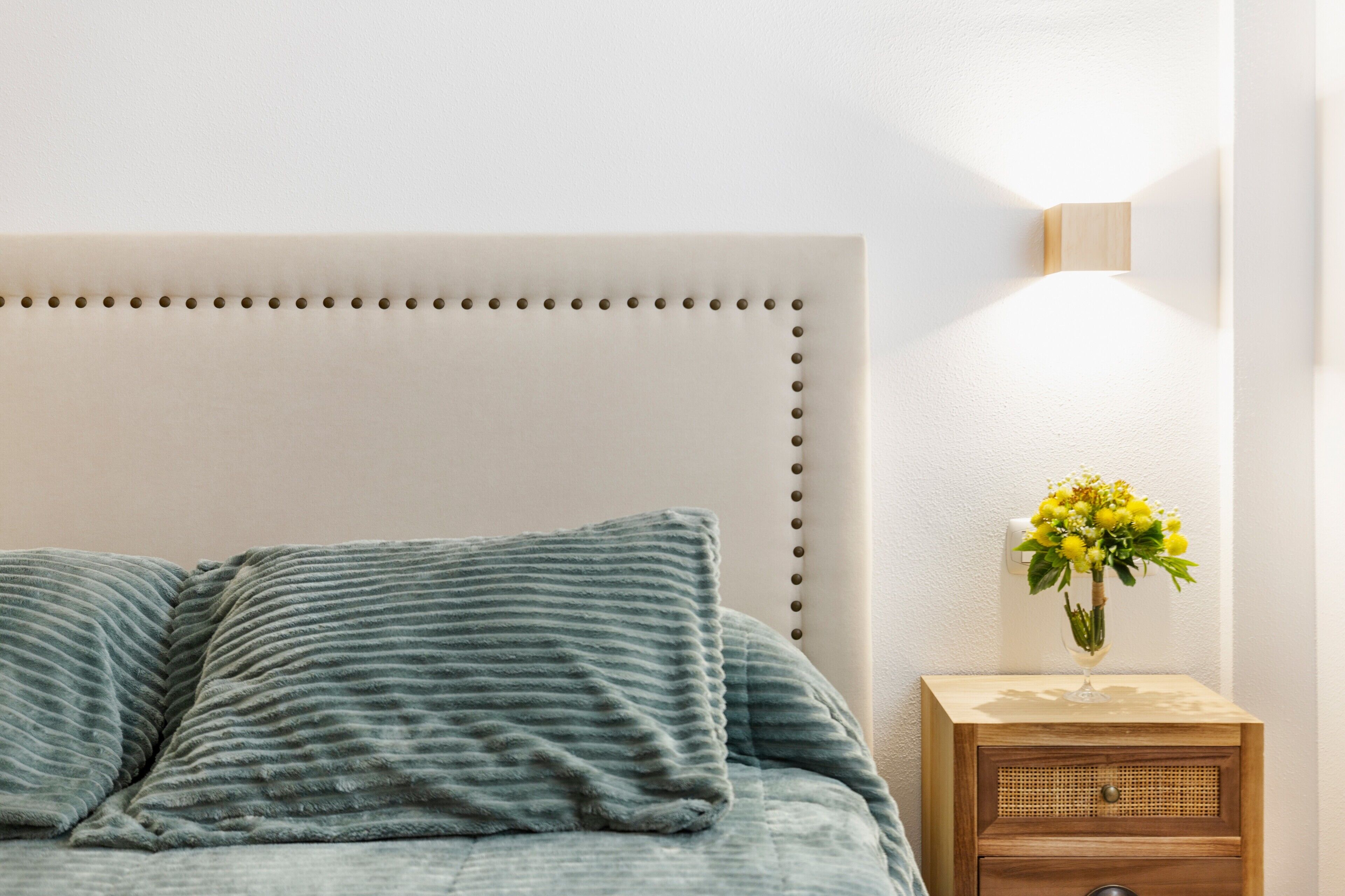 Uma cama com cabeceira, travesseiros, uma mesa de cabeceira com um vaso de flores e um candeeiro fixo na parede.