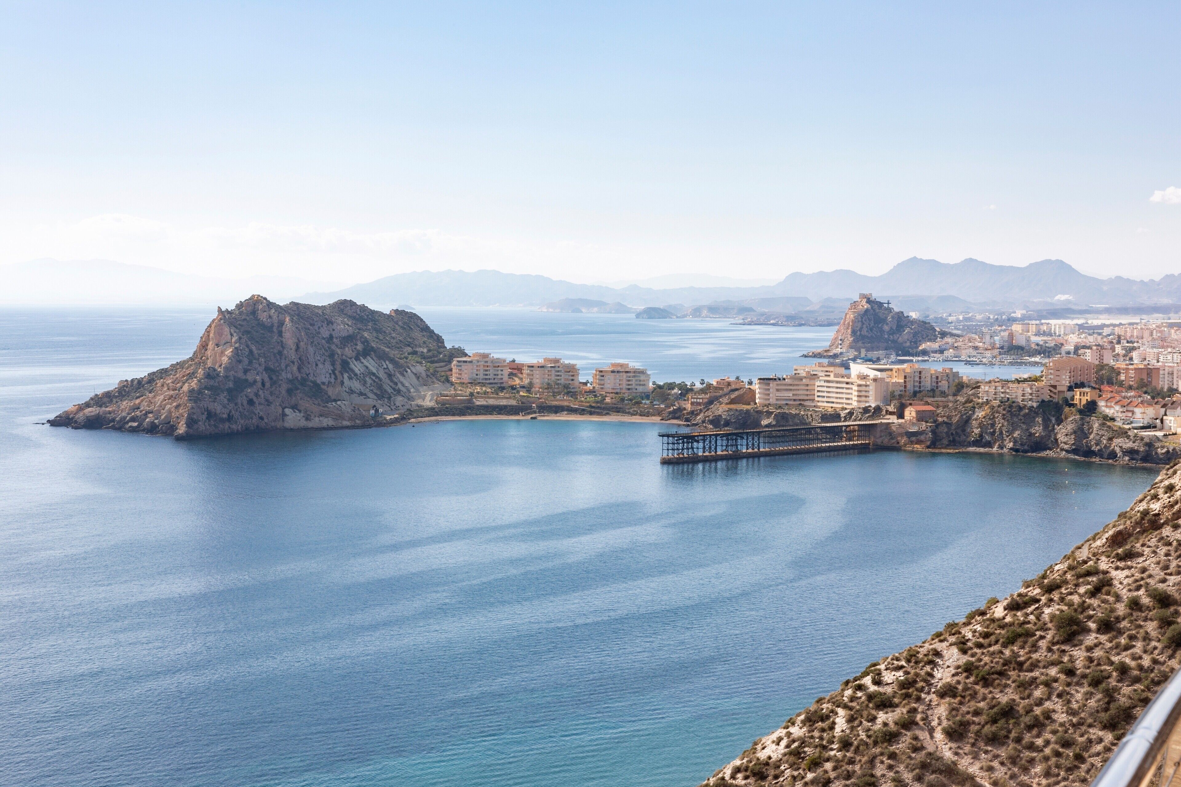 Un paisaje urbano costero con una isla rocosa destacada, un muelle y un puente que conecta con tierra firme.