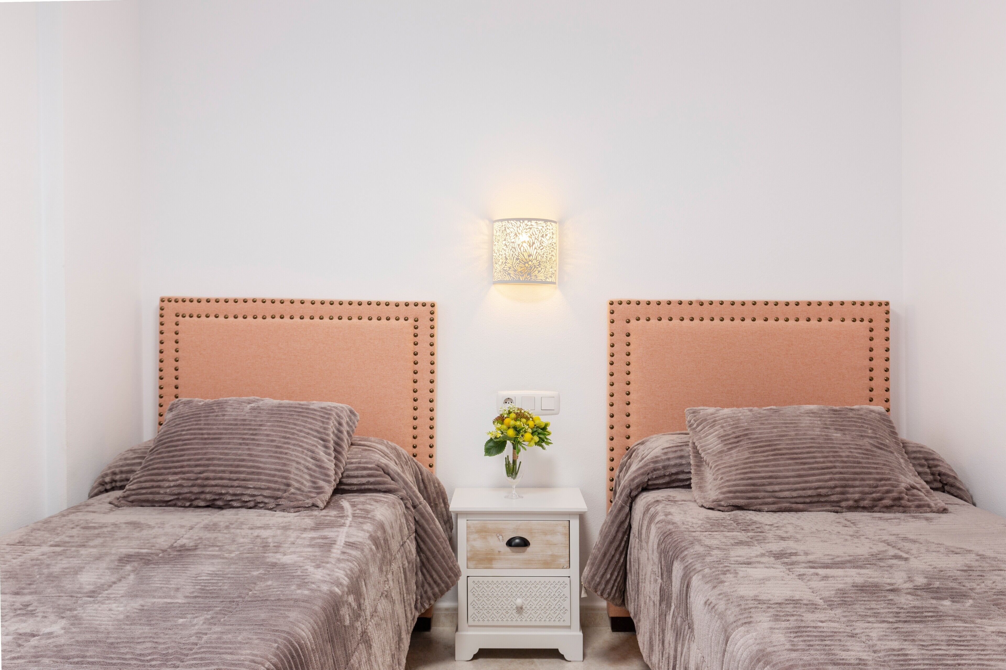 Et soveværelse med to senge, et natbord med en lampe og en vase med blomster.