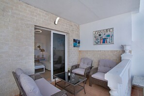 Una stanza con parete in mattoni, mobili in vimini, un tavolino in vetro con una bottiglia e un bicchiere, e un quadro appeso al muro.
