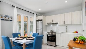 A modern kitchen with white cabinets, a stainless steel refrigerator, a gas stove, and a dining area with blue chairs and a white table.