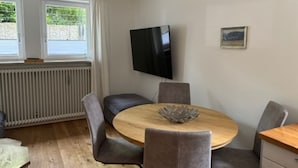 A dining area with a round wooden table, four grey upholstered chairs, a black flat-screen TV mounted on the wall, and a radiator below the windows. - Apartment in the best location in Munich (München)